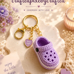 May include: A lavender crochet clog keychain featuring a gold-tone metal ring, a gold-tone lobster clasp, and a small heart-shaped charm with the text "hand made" and a heart symbol. The miniature shoe has a white sole and a black side rivet.