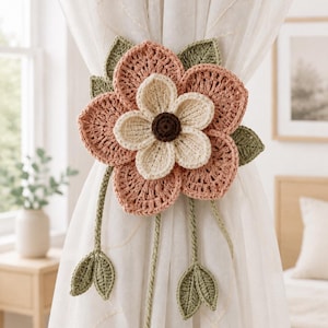 May include: A crochet curtain tieback featuring a large layered flower with dusty pink outer petals, cream inner petals, and a dark brown center. The design includes sage green crocheted leaves and two dangling stems with leaf accents attached to the flower.