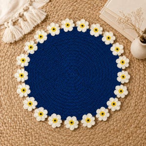 May include: A circular dark blue crochet mat featuring a decorative border of small white flowers with yellow centres. The mat is constructed with a dense, uniform stitch pattern, and the floral accents are evenly spaced around the entire edge.