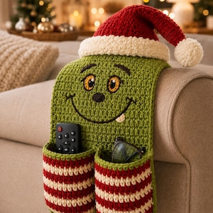 May include: A green crocheted armchair caddy featuring a smiling face and a red and white Santa hat. The caddy has two pockets with red and white horizontal stripes, containing a black remote control and a pair of black-framed eyeglasses.