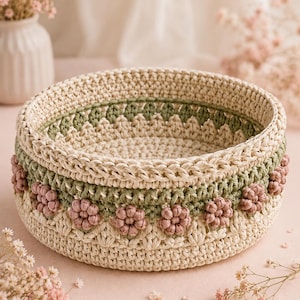 May include: A round, hand-crocheted basket featuring a cream-colored base and rim, a sage green middle band, and a row of small, textured, dusty pink floral embellishments encircling the exterior. The structure is firm with a flat bottom.