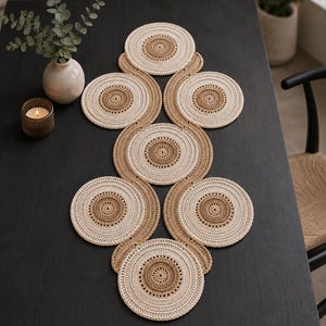 May include: A table runner composed of seven interconnected circular crochet motifs. Each circle features a dark tan centre surrounded by a wider band of cream-coloured yarn. The circles are linked in a vertical, wave-like pattern of alternating colours.