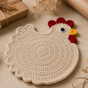 May include: A round, beige crochet coaster shaped like a chicken. The design features a scalloped edge, a red crochet comb and wattle, a yellow beak, and a black and white eye. The body is constructed with a circular stitch pattern in light tan yarn.