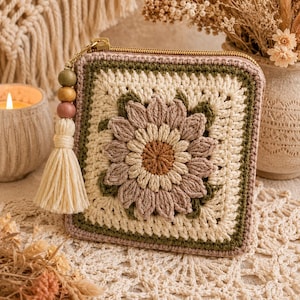 May include: A square crochet pouch featuring a central floral motif in shades of cream, dusty rose, and olive green. The pouch has a gold-tone zipper closure adorned with a tassel made of cream yarn and three wooden beads in green, tan, and rose.