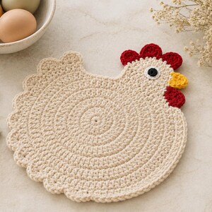 May include: A cream-colored crochet chicken-shaped trivet featuring a circular body, a red comb and wattle, a yellow beak, and a black and white eye. The piece is crafted with a textured stitch pattern and scalloped edges along the tail and body.