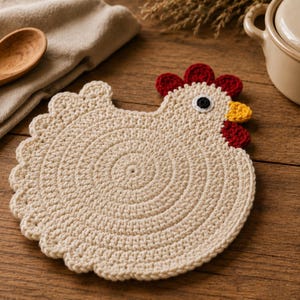 May include: A cream-coloured crochet trivet shaped like a chicken, featuring a circular body with scalloped edges, a red comb, a yellow beak, and a small black eye. The piece is crafted with a dense, textured stitch pattern, creating a flat, round silhouette that mimics the appearance of a hen in profile.