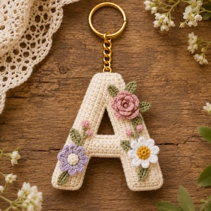 Puede incluir: Un llavero con la letra A tejida a ganchillo en color crema, con un anillo y una cadena de metal dorado. La letra está decorada con pequeñas flores de tela cosidas a mano en tonos rosa, morado y blanco, acompañadas de hojas verdes tejidas.