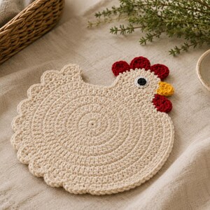 May include: A round, cream-colored crochet coaster shaped like a chicken. The body features a circular stitch pattern with a scalloped edge. It has a red crochet comb and wattle, a yellow beak, and a black and white eye detail on the head.