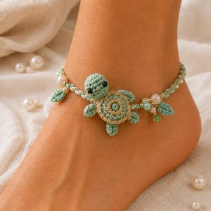 Può includere: Una cavigliera all'uncinetto con un ciondolo centrale a forma di tartaruga marina in filato verde chiaro e beige. Il cinturino è realizzato in corda intrecciata verde chiaro e bianca, decorato con piccole perle bianche, perline verde chiaro e foglie.