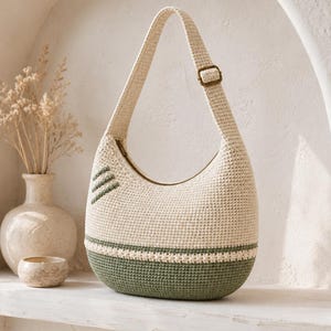 May include: A crescent-shaped crochet shoulder bag in cream and sage green. The bag features a solid cream upper section, a sage green base, and a decorative horizontal band. It includes a single adjustable strap with a bronze-toned metal buckle.