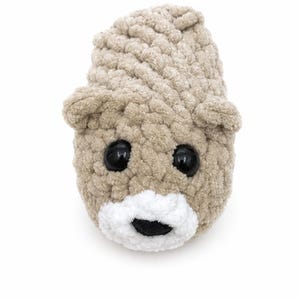 May include: A beige crocheted plush toy shaped like a bear head. The toy features two small rounded ears, two large glossy black plastic eyes, and a white crocheted muzzle area with a small black stitched nose centred on the lower face.
