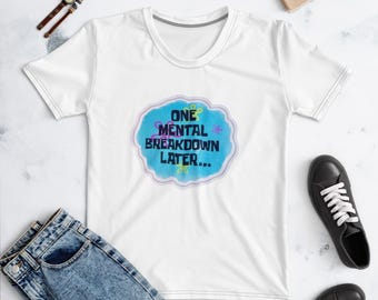May include: A white short-sleeved t-shirt featuring a central blue cloud-shaped graphic with the text "ONE MENTAL BREAKDOWN LATER..." in black, accented by small pink and yellow flower outlines, presented on a flat surface with accessories.