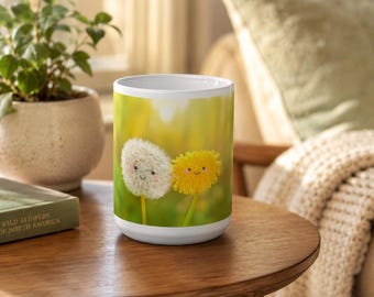May include: A white ceramic mug featuring a printed graphic of two dandelion heads with smiling faces. One dandelion is white and fluffy, while the other is bright yellow. The background of the print shows a soft, out-of-focus green field.