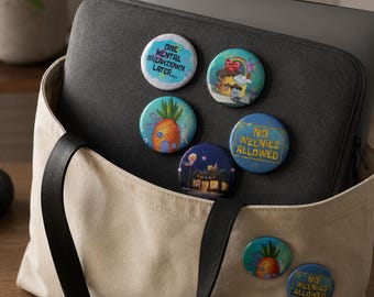 May include: A beige canvas tote bag and a dark gray laptop sleeve featuring seven circular pin-back buttons. The buttons display various colorful cartoon illustrations and text including "ONE MENTAL BREAKDOWN LATER..." and "NO WEENIES ALLOWED."