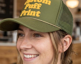 May include: An olive green trucker hat featuring a mesh back and a structured front panel. The front displays the text Custom Puff Print in raised, yellow, rounded lettering. A thin, braided cord runs horizontally across the base of the crown.