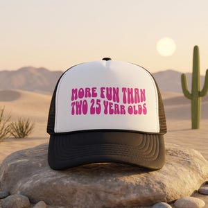 May include: A white and black trucker hat featuring the pink text "MORE FUN THAN TWO 25 YERR OLDS" printed on the front foam panel. The cap has a structured white front, a black brim, and a black mesh back with an adjustable snap closure.