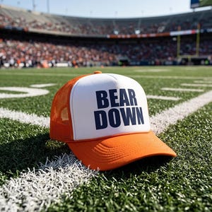 May include: An orange and white trucker hat featuring a mesh back and a solid front panel. The text BEAR DOWN is printed in bold navy blue capital letters on the white front panel. The hat has a curved orange brim and an adjustable snapback closure.
