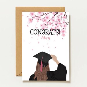Könnte beinhalten: Eine weiße Grußkarte mit einer braunhaarigen Person in schwarzer Abschlussrobe und Doktorhut, darunter rosa Kirschblüten und der Text CONGRATS! Mary. Die Karte liegt auf einem beigen Umschlag und enthält den Text designed by Stud..