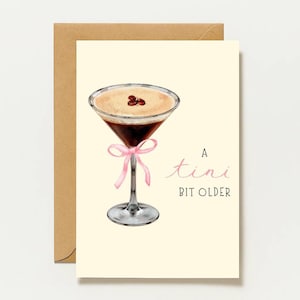 Peut inclure: Une carte de vœux sur fond crème avec une illustration à l'aquarelle d'un verre à martini contenant un cocktail au café, garni de trois grains de café et noué d'un ruban rose. Le texte indique A tini BIT OLDER.