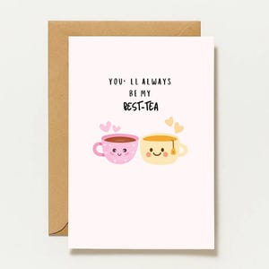 Könnte beinhalten: Eine weiße Grußkarte mit braunem Umschlag, die zwei gezeichnete Teetassen mit Gesichtern zeigt. Die rosa Tasse hat Herzen, die gelbe einen Teebeutel. Der Text lautet: YOU'LL ALWAYS BE MY BEST-TEA. Unten steht: designed by Napel Studio.