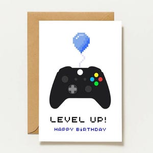 May include: A white greeting card featuring a black pixel art video game controller with a blue pixelated balloon tied to it. Below the controller, the text reads LEVEL UP! HAPPY BIRTHDAY in a pixelated font. The card is placed on a brown envelope.