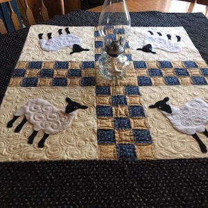 May include: A quilted table runner with a black and white chequered pattern and four white sheep appliques with black faces. The runner is on a black and white patterned table.