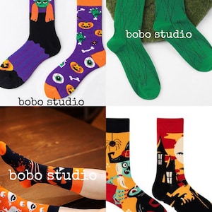 May include: A collection of crew socks featuring various Halloween-themed designs including Frankenstein, pumpkins, eyeballs, bones, ghosts, witches, and haunted houses. The socks display vibrant colors like orange, purple, green, black, and red.
