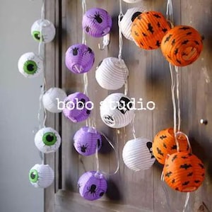 May include: A string of Halloween-themed paper lanterns featuring various designs including green eyeballs, purple lanterns with black spiders, white lanterns with black ghosts, and orange lanterns with black bats, with the text bobo studio visible.