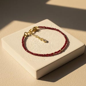 May include: A delicate bracelet featuring small, faceted deep red garnet beads interspersed with smooth gold-toned metal spheres. The piece includes a gold-toned metal chain extender with a small, clear, heart-shaped charm at the end and a clasp.