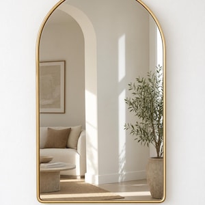 Op de afbeelding: Een boogvormige wandspiegel met een dun, glad, goudkleurig metalen frame. Het glazen oppervlak biedt een heldere reflectie van een binnenruimte, met een witte bank, een ronde salontafel en een potplant, allemaal omlijst door de gebogen bovenkant.