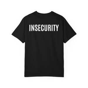以下が含まれることがあります： 背面上部に太字の白いサンセリフ体大文字で「INSECURITY」という単語がプリントされた、黒の半袖Tシャツ。クルーネックで標準的なフィット感があり、無地の黒い生地の質感が特徴的な衣類です。