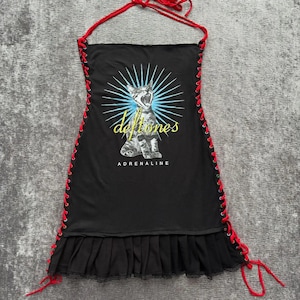 May include: A black halter-style dress featuring red lace-up sides and a ruffled hem. The front displays a graphic of a cat with a blue sunburst background, the yellow text "deftones," and the white text "ADRENALINE" printed below the cat.