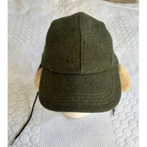 May include: An olive green felt baseball cap featuring a structured crown and a curved brim with visible stitching. The cap includes beige faux shearling ear flaps on the sides and a thin black cord hanging from the left side for adjustment.