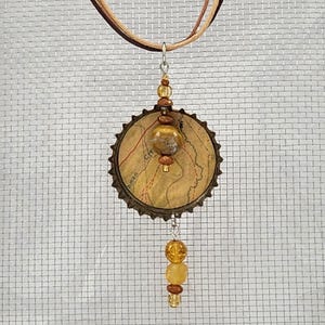 Könnte beinhalten: Eine Anhängerkette mit einer runden Kronkorken-Basis, die mit einem alten Landkartenmotiv bedruckt ist. Das Schmuckstück ist mit bernsteinfarbenen Perlen und einer größeren zentralen Steinperle verziert und hängt an einer mehrsträngigen Kordel.