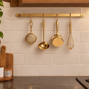 Puede incluir: Un riel de cocina de metal dorado montado horizontalmente, con cuatro utensilios de cocina colgantes: un colador redondo de malla, un cucharón profundo, una cacerola pequeña y un batidor de alambre. Todos tienen un acabado metálico dorado.