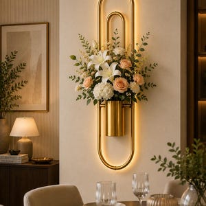 Puede incluir: Un aplique de pared de metal dorado con un marco ovalado vertical e iluminación LED integrada. Un jarrón cilíndrico dorado está fijado en el centro, conteniendo un arreglo de lirios blancos, rosas color melocotón, hortensias y follaje.
