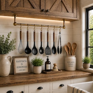 May include: A brass kitchen rail mounted under wooden cabinets holds a set of black silicone utensils with wooden handles, including a whisk, ladle, slotted spoon, pasta server, spatula, solid spoon, and metal tongs, alongside kitchen decor.
