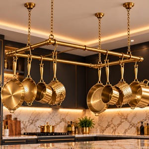 May include: A rectangular brass pot rack suspended from the ceiling by four chains, featuring a pipe-style frame. Eight brass-toned cookware pieces, including various pans and a colander with smooth and hammered textures, hang from S-hooks.