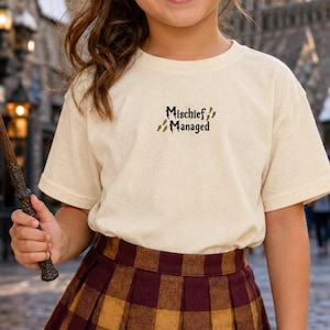 May include: A cream-colored short-sleeve t-shirt featuring the embroidered text "Mischief Managed" in black, accompanied by small brown footprint icons. The shirt is paired with a pleated skirt in a dark red and mustard yellow plaid pattern.