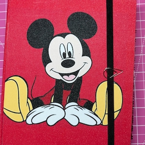 May include: A red notebook featuring a graphic of Mickey Mouse on the front cover. The character is depicted with black ears, a white face, and yellow shoes. A black elastic band wraps vertically around the cover, secured by a silver metal paperclip.