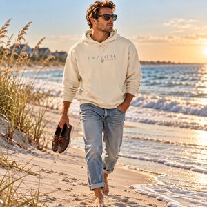 May include: A cream-colored hooded sweatshirt featuring the word EXPLORE printed in black on the chest, paired with light blue denim jeans and dark brown leather boat shoes held in one hand, worn with dark sunglasses.