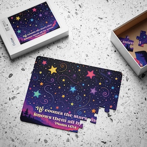 Puede incluir: Un rompecabezas con un cielo nocturno azul oscuro lleno de estrellas coloridas y líneas en espiral. El texto dice, He counts the stars and knows them all by name Psalm 147:4. El rompecabezas se muestra parcialmente armado con su caja blanca.