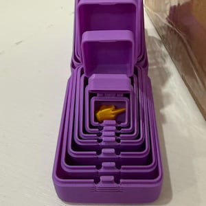 Könnte beinhalten: Ein Set aus ineinander verschachtelten violetten rechteckigen Behältern, die vertikal gestapelt sind. Die Behälter werden zur Mitte hin kleiner, wo sich eine kleine, goldfarbene, dreidimensionale Handfigur mit ausgestrecktem Mittelfinger befindet.