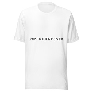 May include: A white short-sleeved t-shirt featuring the black text PAUSE BUTTON PRESSED centered on the chest. The garment has a classic crew neckline and a straight hemline, providing a clean and simple look for casual everyday wear.