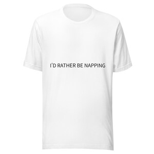 May include: A white short-sleeved t-shirt featuring the black text I'D RATHER BE NAPPING printed across the center of the chest. The garment has a standard crew neckline and a straight hemline, providing a simple and clean appearance for daily wear.
