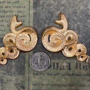 May include: Two gold-colored snake-shaped decorative objects. Each snake is coiled with a detailed scale pattern and a raised head. A US quarter is placed between the objects for size comparison. The background is a vintage newspaper.