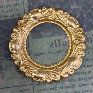 May include: A gold-colored, ornate circular frame with a detailed, vintage design. The frame features a central opening and is decorated with floral and scrollwork patterns, suitable for crafts and decorative projects.