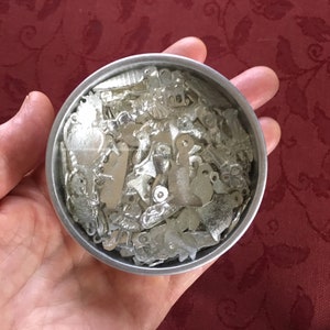 May include: A small, round, silver-colored tin filled with numerous small, silver-toned metal charms. The charms are various shapes, including hearts, keys, and other decorative designs. The tin is held in a person's hand, with a red background.