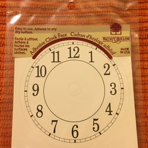 May include: A white adhesive clock face with black numbers 1 through 12. The clock face has a red adhesive border with the text "Adhesive Clock Face" and "Cadran d'horloge adhésif". The text "Walnut Hollow" and "Made in USA" is printed on the right side of the clock face.