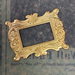 May include: Ornate gold-colored metal frame with intricate floral and scrollwork details. The rectangular frame has a central opening and is likely for a photograph or artwork. The frame is set against a blue-toned background with faint text.